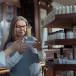 Elderly woman receiving assistance from a pharmacist in a pharmacy with product shelves.