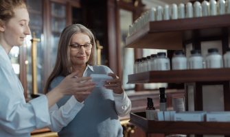 Elderly woman receiving assistance from a pharmacist in a pharmacy with product shelves.