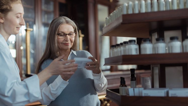 Elderly woman receiving assistance from a pharmacist in a pharmacy with product shelves.
