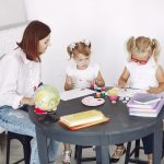 Small kids attentively painting in kindergarten with adult assistance while sitting together at round table