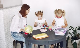 Small kids attentively painting in kindergarten with adult assistance while sitting together at round table