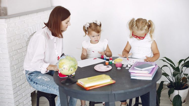 Small kids attentively painting in kindergarten with adult assistance while sitting together at round table