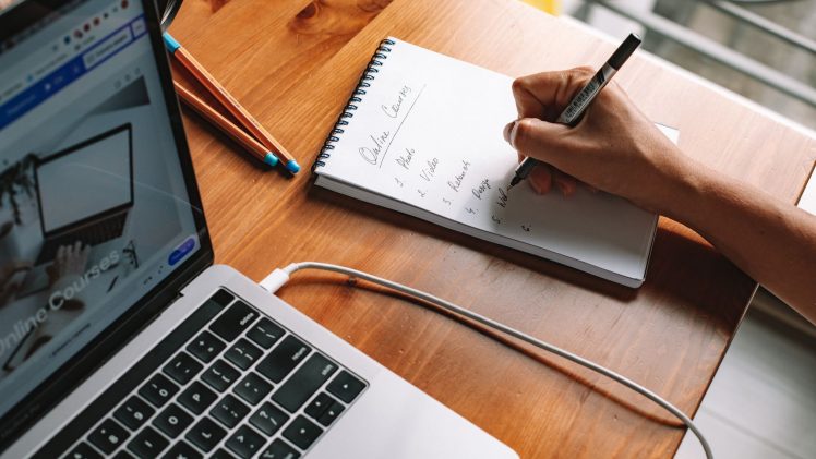 A person writing notes while researching online courses on a laptop in a home setting.