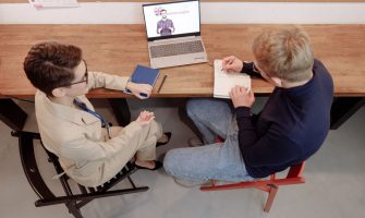 Two adults engaged in online learning, one taking notes during an English study session.