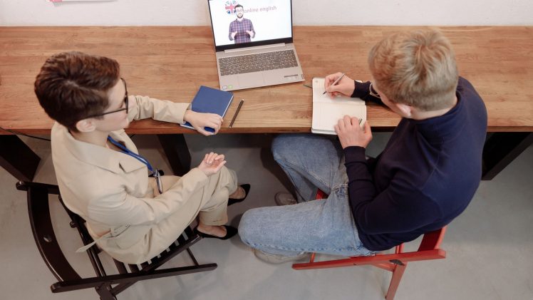 Two adults engaged in online learning, one taking notes during an English study session.
