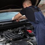 A mechanic in blue overalls works on a car engine outdoors, showcasing vehicle maintenance skills.