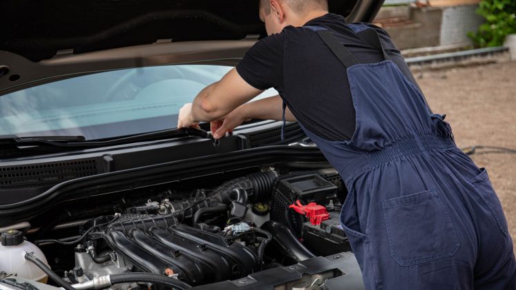A mechanic in blue overalls works on a car engine outdoors, showcasing vehicle maintenance skills.