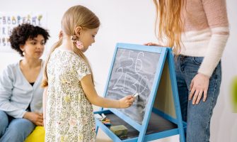 A young girl writes numbers on a chalkboard in a classroom, supervised by adults.