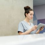 A receptionist in scrubs using a tablet at an office reception desk.