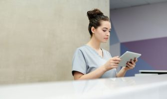 A receptionist in scrubs using a tablet at an office reception desk.
