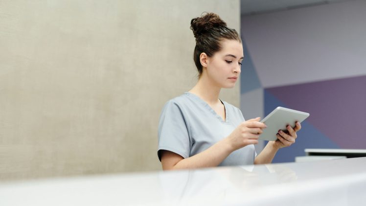 A receptionist in scrubs using a tablet at an office reception desk.