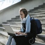 Woman using laptop on outdoor stairs, studying online with headphones and backpack.