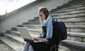 Woman using laptop on outdoor stairs, studying online with headphones and backpack.