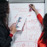 Two professional women discuss and strategize on business development in a modern office setting.