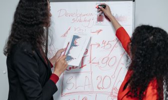 Two professional women discuss and strategize on business development in a modern office setting.