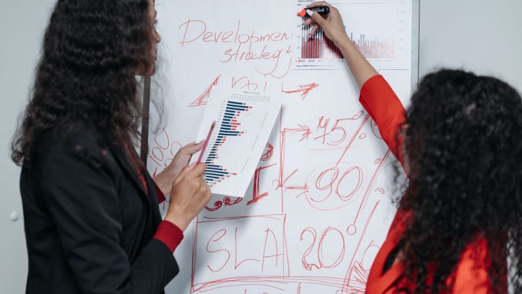 Two professional women discuss and strategize on business development in a modern office setting.