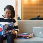 A woman studying Java programming on a sofa with a MacBook and other computer books nearby.