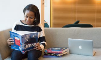 A woman studying Java programming on a sofa with a MacBook and other computer books nearby.