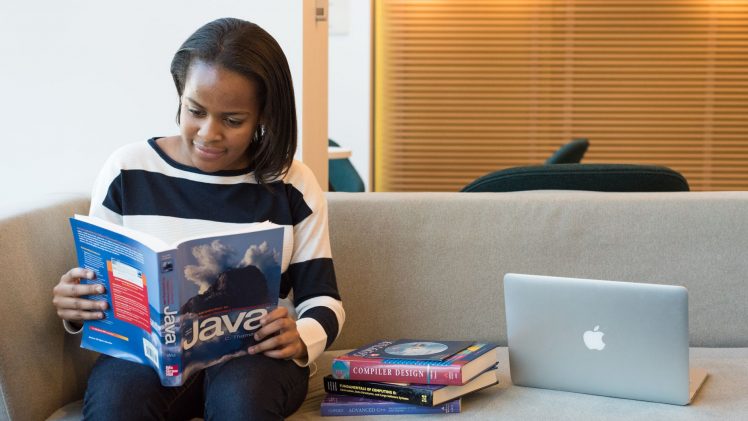 A woman studying Java programming on a sofa with a MacBook and other computer books nearby.