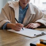 Crop young female unemployed job seeker filling questionnaire sitting at table during interview in office