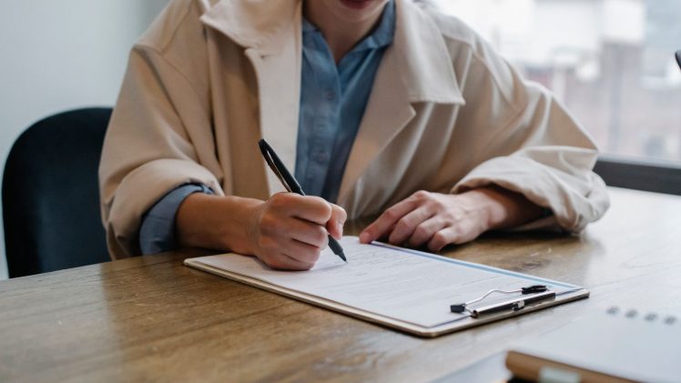 Crop young female unemployed job seeker filling questionnaire sitting at table during interview in office