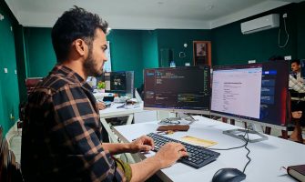 A software developer working on code at a dual monitor setup in a modern office.