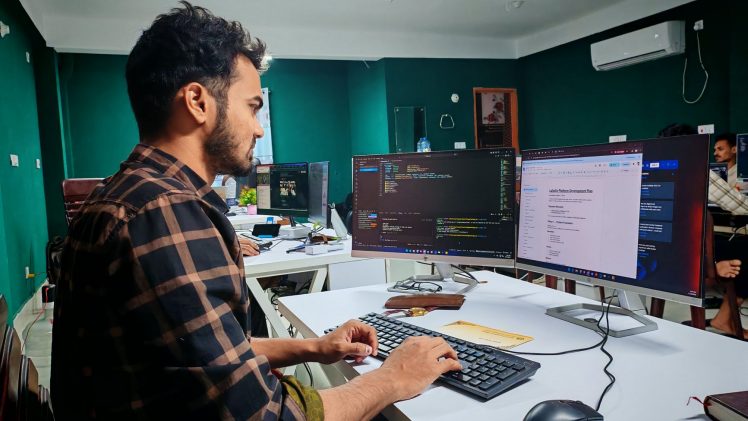A software developer working on code at a dual monitor setup in a modern office.