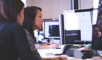 Two women working together on code at a computer in a modern office setting.