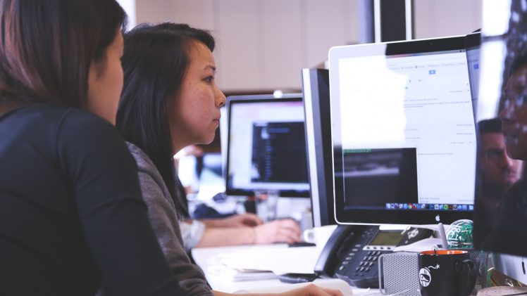 Two women working together on code at a computer in a modern office setting.