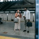 Two professionals chatting at a modern train station entrance, holding coffee and bags.