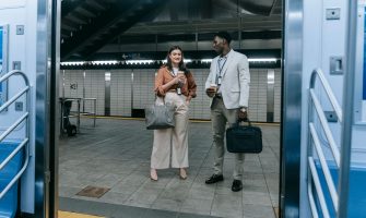 Two professionals chatting at a modern train station entrance, holding coffee and bags.