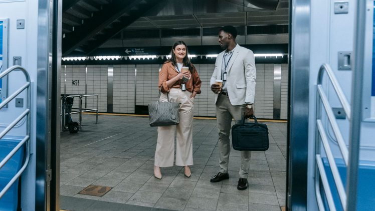 Two professionals chatting at a modern train station entrance, holding coffee and bags.