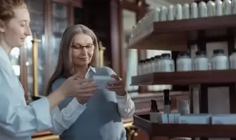 Elderly woman receiving assistance from a pharmacist in a pharmacy with product shelves.