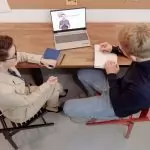Two adults engaged in online learning, one taking notes during an English study session.