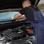A mechanic in blue overalls works on a car engine outdoors, showcasing vehicle maintenance skills.