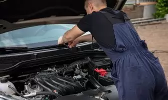 A mechanic in blue overalls works on a car engine outdoors, showcasing vehicle maintenance skills.