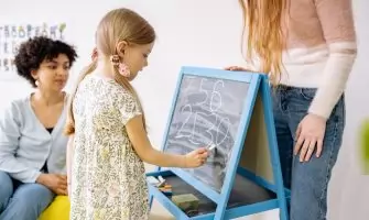A young girl writes numbers on a chalkboard in a classroom, supervised by adults.