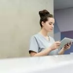 A receptionist in scrubs using a tablet at an office reception desk.