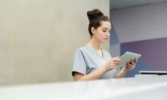 A receptionist in scrubs using a tablet at an office reception desk.