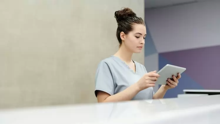 A receptionist in scrubs using a tablet at an office reception desk.