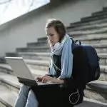 Woman using laptop on outdoor stairs, studying online with headphones and backpack.