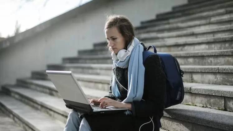 Woman using laptop on outdoor stairs, studying online with headphones and backpack.