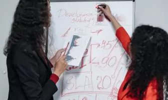 Two professional women discuss and strategize on business development in a modern office setting.