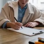 Crop young female unemployed job seeker filling questionnaire sitting at table during interview in office