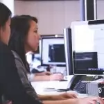Two women working together on code at a computer in a modern office setting.