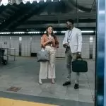Two professionals chatting at a modern train station entrance, holding coffee and bags.